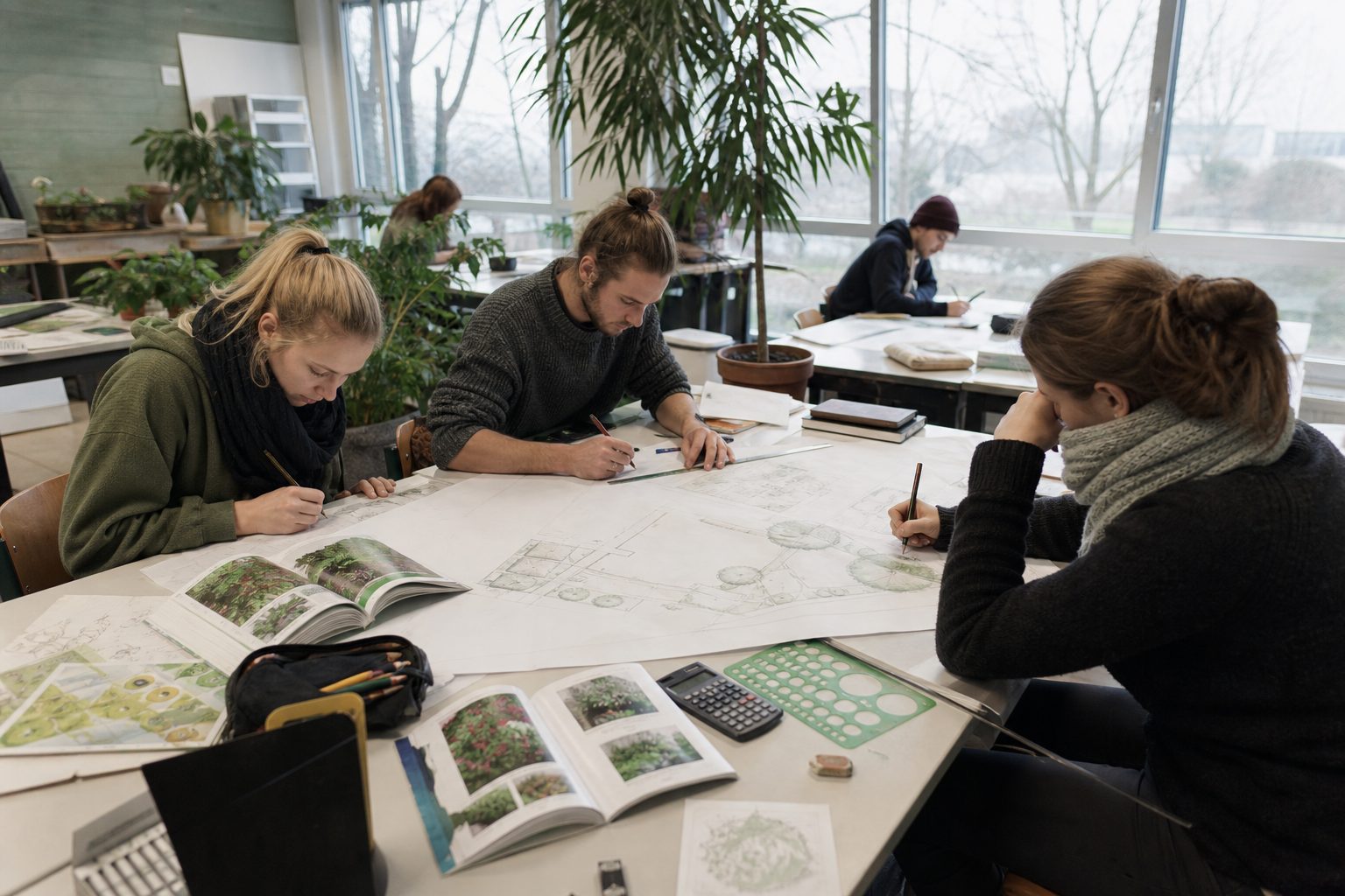 Meisterschüler zeichnen einen Gartenplan im Unterrichtsraum einer Fachschule, Pflanzenkataloge auf dem Tisch