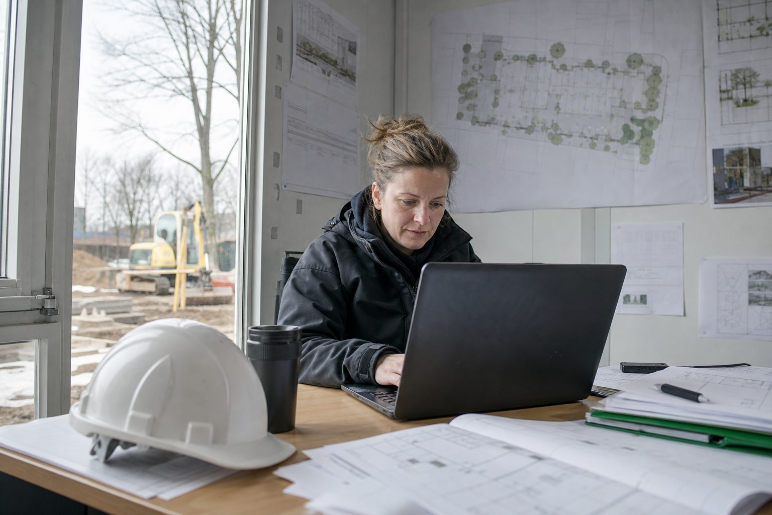 Bauleiterin prüft Ausschreibungsunterlagen am Laptop im Baucontainer, Pläne an der Wand